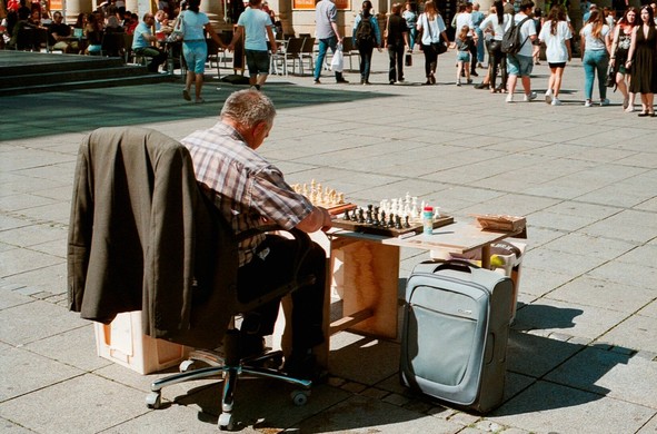 A Chess player waiting for opponents on the street. Pedestrians are passing by