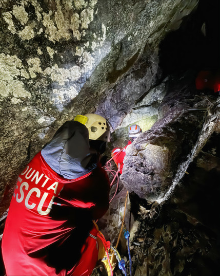 The image shows a dramatic cave rescue scene inside a dark, rugged cavern. Two rescue team members, dressed in red Mountain Rescue jackets and helmets, are assisting a caver in navigating a steep, narrow rock passage. The rescuer in the foreground, with "RESCUE" partially visible on their back, is wearing a helmet with a headlamp, while another rescuer ahead is securing ropes and guiding the exhausted caver upwards. The cave walls are rough and damp, illuminated by the team’s lights, casting shadows on the jagged rocks. The scene is intense, highlighting the physical challenge and skill required for such a rescue.