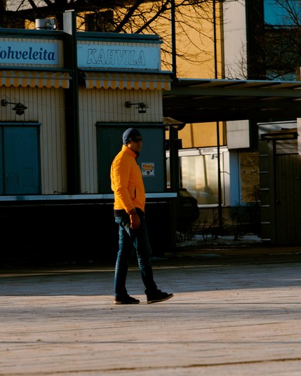 The image shows a person in a bright orange jacket and dark pants walking across a paved area. They are wearing a beanie and sunglasses. In the background, there is a building labeled "KAHVILA," suggesting it might be a café or kiosk. The overall setting appears to be urban, with the presence of buildings and trees. The lighting suggests it's daytime, likely in the afternoon with long shadows.