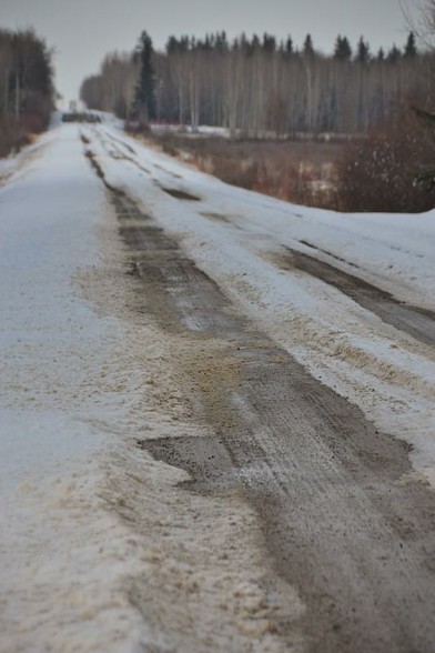 Looking up a gravel country road. There is mud-splashed snow on both sides and centre with double tire tracks that are mostly clear of snow,  with puddles, mud and slush. In the distance we see wetland and woodland to the sides of the road. The day is overcast.