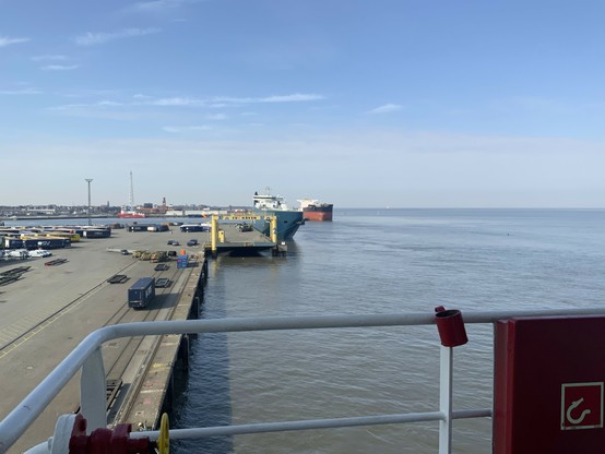 Hafen mit Kaianlagen und Schiffen. Im Vordergrund: Reeling mit einem roten Schlauchkasten