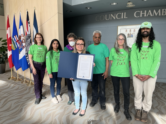 Senator Simons, in a green blazer, purple scarf and N95 mask, stands with a group of soil advocates wearing bright green T-shirts that read Save our Soil, outside Edmonton City Council Chambers. In the middle of the group, a City clerk holds open a big blue folder with the soil health proclamation.