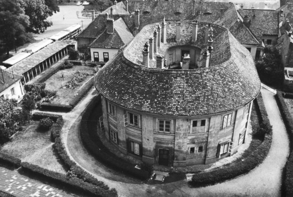 A aerial view of a two storied stadium shaped building. That is it is curved at each end but is straight in the middle section. There are many chimneys on the roof. The building is surrounded a garden on one side and various other buildings as well.