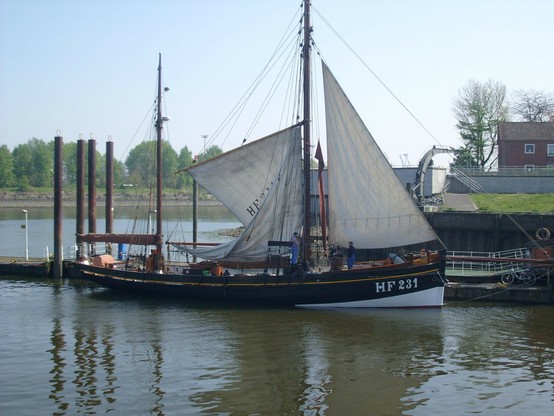 Besegelter Fischkutter im Hafen. Schwarzer Rumpf, weißes Unterwasserschiff. Das Schiff liegt an der Pier, das Focksegel ist ganz, das Großsegel teilweise zur Überprüfung gesetzt.