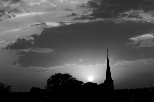 SchwarzundWeiß-Aufnahne 
Im Vordergrund die Silhouette von Bäumen und des Dorfkirchiens. Im Hintergrund die tiefstehende Sonne mit einem gemischten Himmel mit Wolken und unterschiedlicher Farbgebung 😉🤗
Matze-work