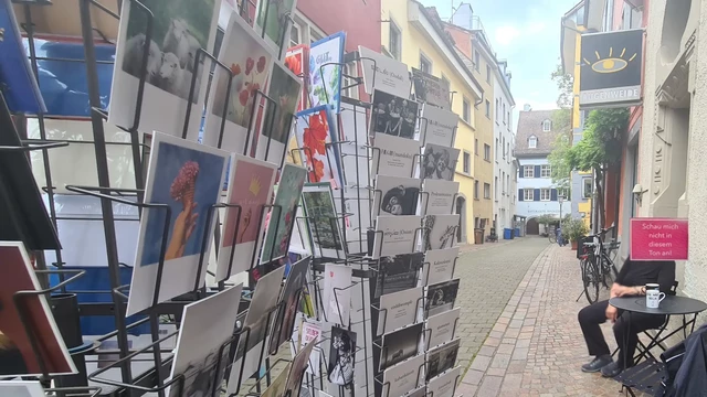 Altstadtgasse mit teils Kopfsteinpflaster, links Postkartenständer mit Sprüchekarten, rechts große Karte mit "Schau mich nicht in diesem Ton an" überm Kopf einer Person die an einem kleinen Bistrotisch mit Kaffee sitzt