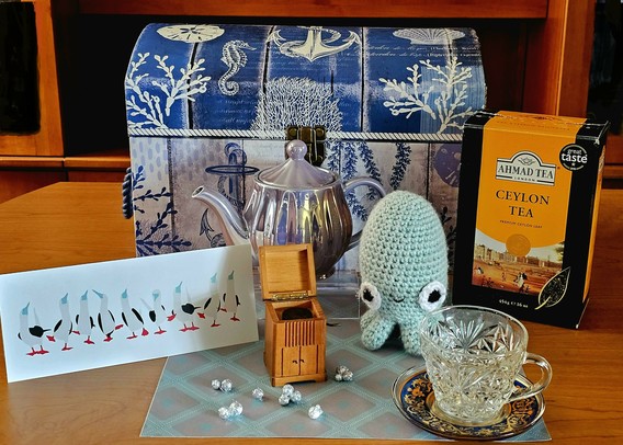 Whimsical tea tableau in an octopus's formal parlour with a gramophone, cut glass tableware, gleaming opalescent teapot, diamond patterned rug, an adorable crocheted octopus, a line of dancing seabirds, sparkly silver bubbles, and Ceylon black tea from Ahmad. The backdrop is a small trunk with an aquatic design.