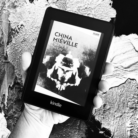 A hand holds a Kindle displaying the book cover of "The City & The City" by China Miéville. The background features a textured, monochromatic surface.
