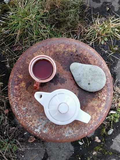 A tea set consisting of rustic teapot and cup on a rusty metal plate, accompanied by a  beautiful smooth gray pebble.