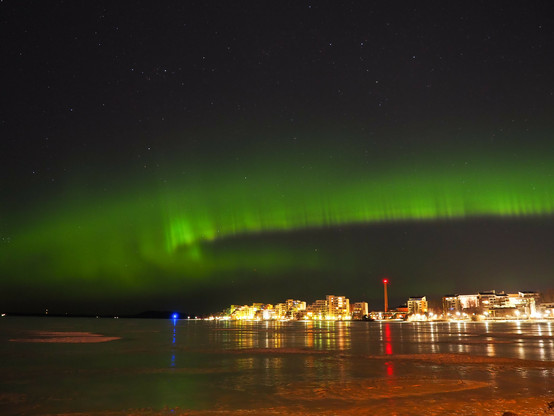 Picture of green northern lights going across the starry sky above a city. Picture is taken at the shore of a frozen lake so there is some reflection from the city lights and auroras on the ice.