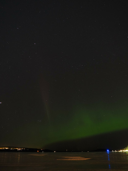 Picture of green northern lights going across the starry sky above a frozen lake. On the left side there is one faint red line of aurora's going straight up. Ice reflects some city lights across the lake.