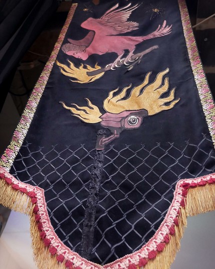 Grand étendard noir vertical de forme médievale, avec pointe en bas ornementée de broderies et de franges dorées. En couture et en broderie, Il représente une harpie (créature mi-femme mi-oiseau souvent utilisée comme insulte envers les femmes en colère). Cette créature volante tient une torche enflammée avec laquelle elle a mis le feu à une caméra de surveillance située sous elle, qui dépasse d'une clôture en grillage. Au dessus d'elle scintillent les broderies dorées de deux étoiles contenant les lettres E (pour égalité) et A (pour Anarchie). Cet étendard veut symboliser la lutte contre l'Etat sécuritaire et l'enfermement.