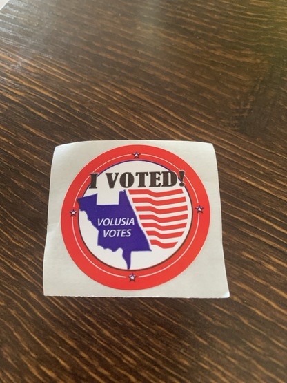 A photo of a sticker I received when I early voted today.  Red circle with an American flag inside  that says “Volusia Votes”
