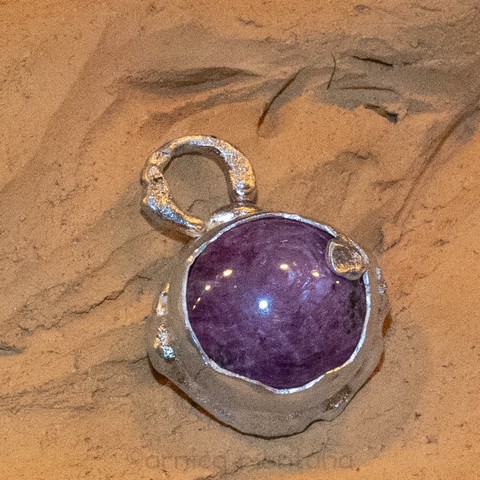 A round pendant featuring a large purple gemstone set in a textured silver frame, resting on a pottery surface.