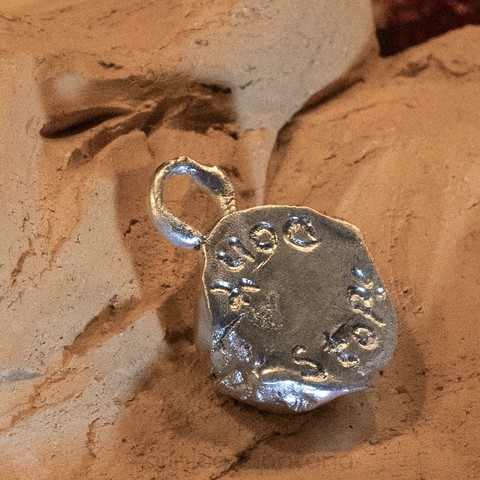 A silver pendant with an irregular shape and a loop for attachment, resting on a textured, neutral-colored surface. The pendant features the inscription: Don’t Stop!