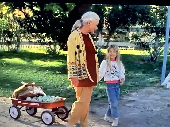 An elderly woman wearing a colorful sweater and a young girl are walking together outdoors. The girl is smiling and holding the woman's hand. A small wagon pulled by the woman contains a cow and some green objects, set against a background of grass and trees.