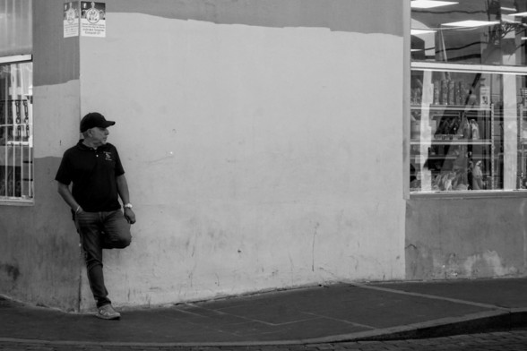 Black and white photo of a man leaning against a blank wall, one foot on the wall behind him. To the far right of the frame is a shop window, its wares barely visible through the glass.