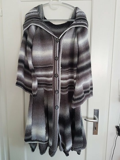 A long, flowing cardigan crafted with Tunisian crochet hangs on a white door. The cardigan features a hood and a gradient pattern transitioning through shades of gray, black, and white. Its design includes a button-up front with five buttons, pockets, and a loose, draping style at the bottom