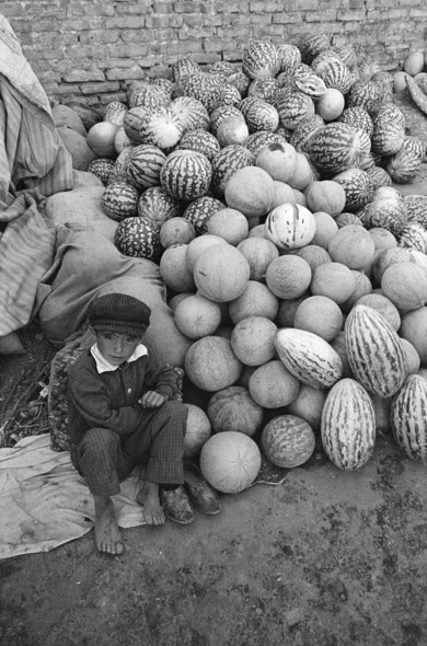 A black and white photo of a young, barefooted boy sitting on the ground next to a large pile of various melons. He is looking up at the camera. Behind the melons is a brick wall.