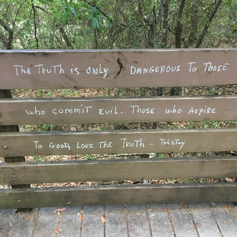 A partial view of a pedestrian walkway through the woods.  Focus of the shot are the path's side railings, consisting of 5 horizontal, parallel boards with spacing between, painted light brown and supported by vertical posts.  Upon the top three boards are written (in white chalk) "The truth is only dangerous to those who commit evil.  Those who aspire to good, love the truth.  -Tolstoy"