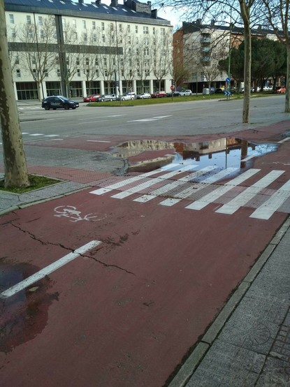 Foto del mismo tramo del carril bici de la foto anterior, esta vez hecha mas cerca de un súper charco que justo está en un cruce de dos carriles bicis y que ocupa todo el carril, haciendo que las bicis tengan que invadir la acera si no quieren pasar  por encima
