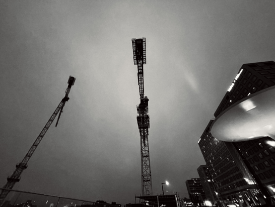 Silhouetted construction cranes tower against a moody gray sky, framed by modern buildings in a cityscape.