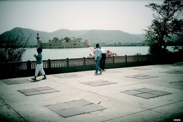 Dunkel getöntes Farbfoto, aufgenommen in Jaipur, Rajasthan, Indien. Blick auf den Wasserpalast Jal Mahal im Man Sagar-See. Im Vordergrund ein gefliester Platz mit ein paar Leuten, begrenzt von einer Balustrade, hinter der Büsche und ein Baum wachsen. Dann der See, das Schloss, die Berge und der diesige Himmel.