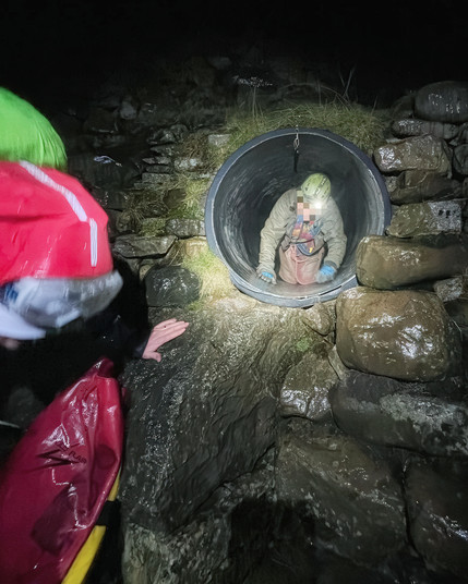 A person in a helmet, their face blurred for privacy, is emerging from a large circular pipe in the dark, wet environment of a rocky hillside, with a rescuer wearing a bright red jacket and a green hood reaching out to assist. The person is wearing an oversuit and gloves, crawling forwards while water glistens on the surrounding stones and the rescue equipment.