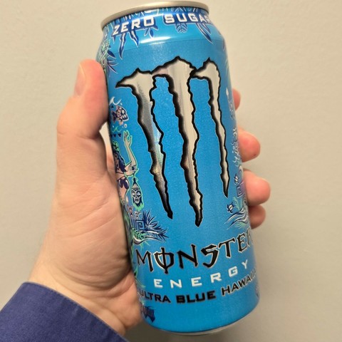Photograph of  white person's hand holding a blue can of Monster Energy that says "Ultra Blue Hawaiian" at the bottom. Centralized is a silvery Monster logo (a large M that looks like three claw marks).