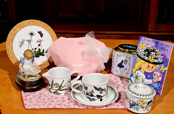 A birthday tea tableau in honor of a friend. There is Portmeirion Botanic Garden china, a bisque porcelain music box statuette of a little girl reading a book, a frilly pink cozy cover and pink calico tea towel, and a birthday card from the 1950s. The tea is Alice In Wonderland tea from Harney and Sons.