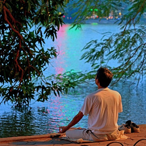 Photo shows a man in white shirt sitting in meditation pose on the low wall surrounding Hoan Kiem city lake. In the background is the lake water disturbed by evening breeze, and in the background on the other side of the lake there are bokeh-typical shapes of colourful lights and buildings. The mans position is framed by low hanging green branches of trees on both sides. He sits on some sort of lightly coloured plastic mat, and his sneakers are placed neatly together to the right. It is a photo taken just after sunset, and the artificial light on the man is soft warm, while the background is a colder blue hue.