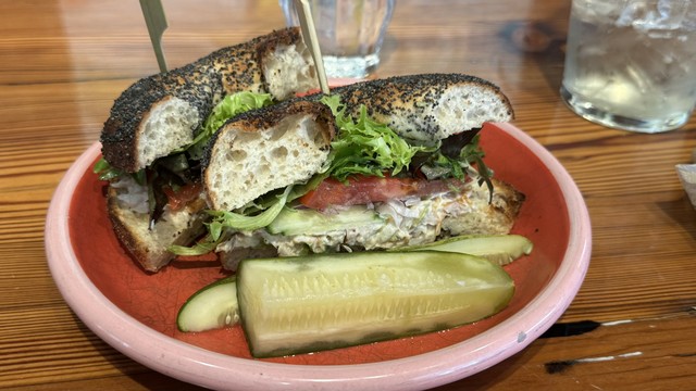 A poppyseed bialy with lettuce, tomato, and whitefish salad, on a pink and red plate with a pickle spear.