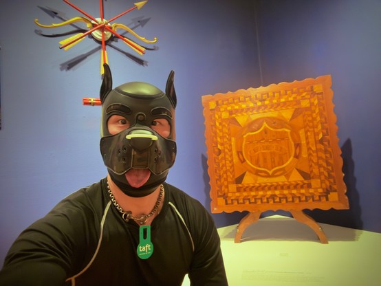 A captivating museum selfie featuring an enthusiast in a detailed black leather puppy hood with prominent pointed ears and an expressive tongue playfully sticking out. They're wearing a sleek black fitted top with white piping trim and a metallic chain collar with a green "taft" tag. Posed beside them is a striking piece of historical woodwork - the Independent Order of Odd Fellows Marquetry Table from 1933. The intricately crafted wooden display features geometric patterns with contrasting light and dark woods forming elaborate borders surrounding a central shield-like emblem. Fascinatingly, the central design resembles a round-faced canine with what appears to be an open mouth and collar chain, creating a serendipitous visual harmony with the puppy hood enthusiast posing alongside it. Above in the background hangs a decorative starburst installation with red and golden arrows. The rich amber tones of the marquetry work beautifully against the purple-blue museum wall. #PuppyPlay #LeatherCommunity #HistoricalArtifacts #OddFellows #MarquetryArt #MuseumVisit #WoodworkArt #FraternalHistory #MuseumMoments #CreativeConnections #BlueskyBuzz #MuseumAdventures  #PupPlayCommunity #PuppyPlayCommunity #PuppyPride #PupPride #mueseumselfie