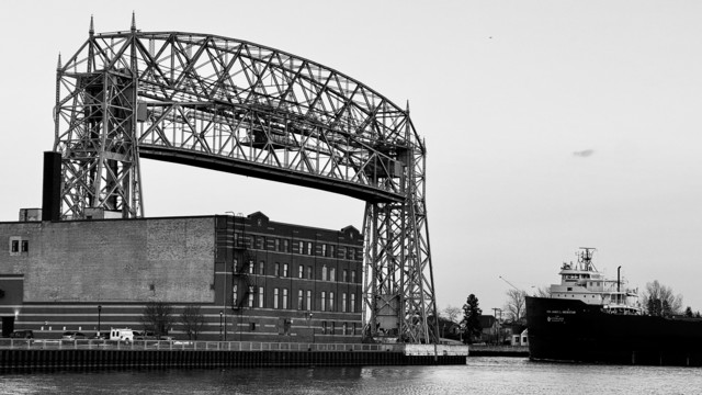 A black-and-white image featuring a large, steel lift bridge above a waterfront; and outbound freighter approaches soon to pas below the bridge. A historic brick building is visible on the left side of the image providing scale for both the bridge and the imminsity of the laker. The scene conveys an industrial atmosphere with a cloudy sky.

The laker is the Hon. James L Oberstar