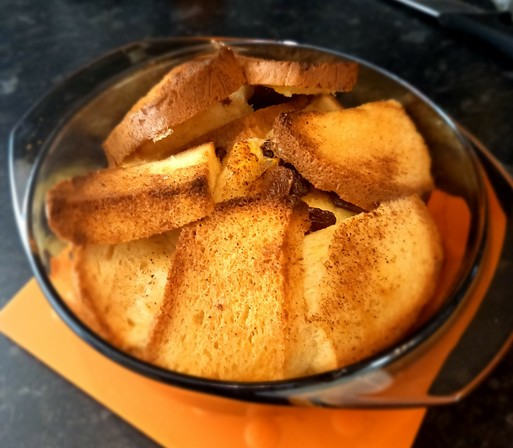 A round dish made of smoked glass standing on a bright orange heat proof mat. The dish is full of layers of small slices of white bread. The lower half is set as a thick bready custard. The top layer is crisp browned bread.
The smell is delicious.