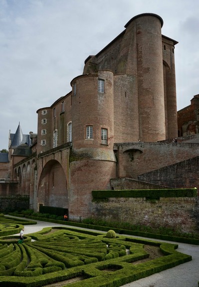 Vue en contre plongée d’une construction ancienne en brique rose massive rappelant un peu une forteresse du Moyen-Âge. Au pied de celle-ci, des buis taillés avec art dessinent des motifs sophistiqués.