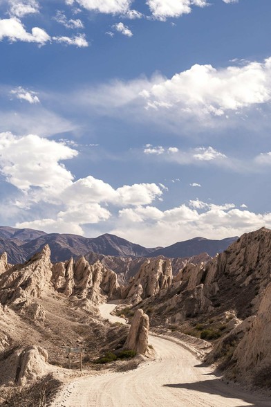 A desert landscape with a dirt road winding through rocky formations.

These formations have sharp, jagged peaks, and the scene is set against a backdrop of mountains in the distance.

The sky above is blue with scattered clouds, creating a striking and dramatic landscape.