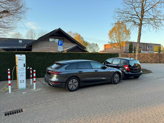 Auf dem Foto ist eine öffentliche E-Ladesäule zu sehen, an der ein Elektroauto (ein dunkelgrauer Limousinen-Kombi) ordnungsgemäß angeschlossen und am Laden ist. Direkt hinter dem Elektrofahrzeug parkt ein schwarzer Mercedes-Benz (Verbrenner), der das E-Auto blockiert und somit das Ausparken erheblich erschwert oder unmöglich macht. Der Parkplatz ist deutlich mit einem Schild als Ladezone für Elektrofahrzeuge gekennzeichnet. Das Verhalten des Benz-Fahrers ist rücksichtslos und behindert sowohl den Ladevorgang als auch die Weiterfahrt des E-Autos.