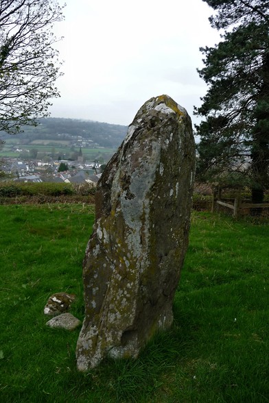 An upright slab of stone, over 6 feet tall, standing in a grass field with trees on either side. The setting is elevated over a valley, and the houses and church spire of small town can be seen in the valley below. Wooded hills rise beyond that. It's a bit grey and overcast.
