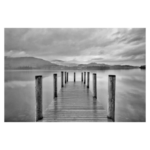 This is a black and white landscape format image of a jetty pointing out across a lake. The viewer looks straight down the jetty into the centre of the image. In the distance mountains are shrouded in clouds. The water of the lake reflects the mountains and the clouds. Taken in the Lake District with a Sony RX100.
