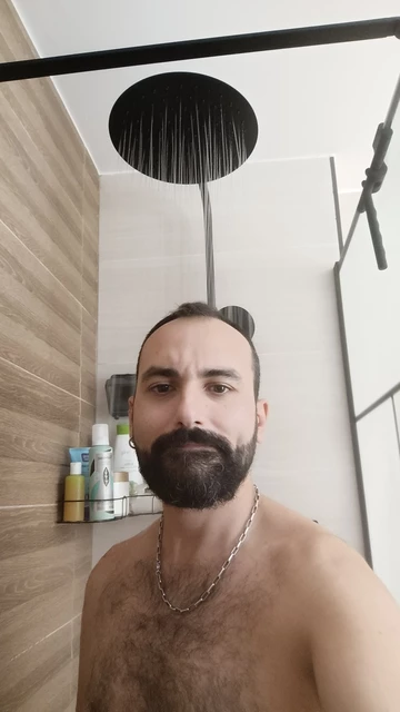 Yo tomándome un selfie en la ducha. Llevo una cadena plateada en el cuello. El agua cae desde una alcachofa de ducha negra en el techo, y al fondo se ven productos de cuidado personal organizados en una repisa.