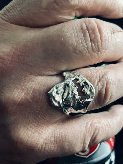A hand is displaying a unique silver ring with an organic, textured design. The ring appears to have a hollow or cut-out section, and the hand is partially holding a can, suggesting a casual setting.