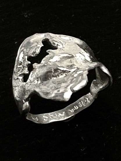A textured, abstract silver ring with organic shapes and cutouts, resting on a dark background. Inscription: stay wild!