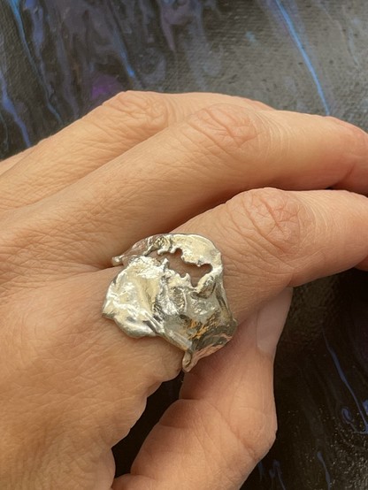 A hand is shown wearing a unique, textured silver ring that has an organic, abstract shape. The background features a dark, glossy surface.