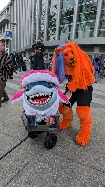 Zander, the orange lion with an orange mane with a blue stripe, standing next to trans icon Jeff the Land Shark from Marvel, wearing a pink and white shark suit