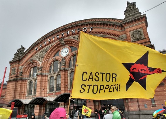 Fahne Castor Stoppen mit Anti-Atom Sonne mit Mundtuch vor schwarzem Stern weht vor dem Gebäude des Bremer Hbf. Personen stehen davor.