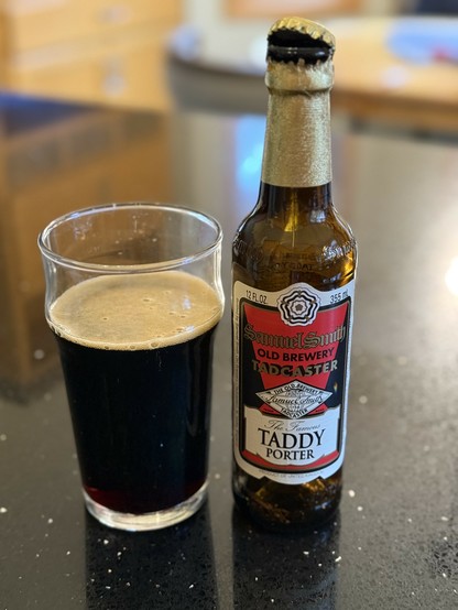 A pint glass full of dark beer next to a brown bottle with a wrinkly gold neck foil and a white label with a red and gold background.
