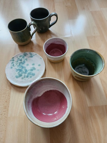 Das Bild zeigt Schalen, Becher, Tassen und einen Teller auf einem Holzuntergrund. Die Keramiken sind Beerenfarben, weiß, türkis und blau glasiert.