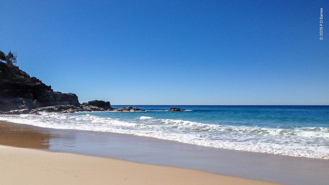 Beach scene with a clear blue sky and a rocky headland coming down into the sea on the left. The sea is calm, a deep blue on the horizon, a lighter blue closer to shore, and just a small line of foam where small waves are finally breaking on the golden sand.