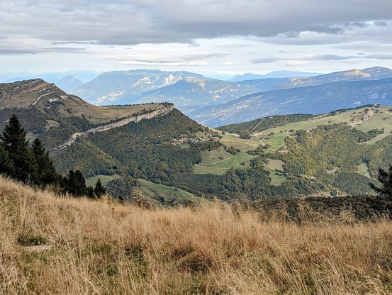 A wide, slightly high-angle shot captures a vast mountainous landscape under an overcast sky. In the foreground, dry, golden-brown grasses dominate, slightly blurred. Beyond this, a series of rolling hills and mountains stretch into the distance. The closest mountains are covered in patches of green vegetation and darker evergreen trees. A distinctive, long ridge with a visible path or road running along its top cuts across the mid-ground. Further back, layers of progressively lighter blue-grey mountains fade into the horizon, some with hints of snow on their higher peaks. The sky is a uniform grey, suggesting a cloudy day. The overall impression is one of expansive natural beauty and a sense of remoteness.